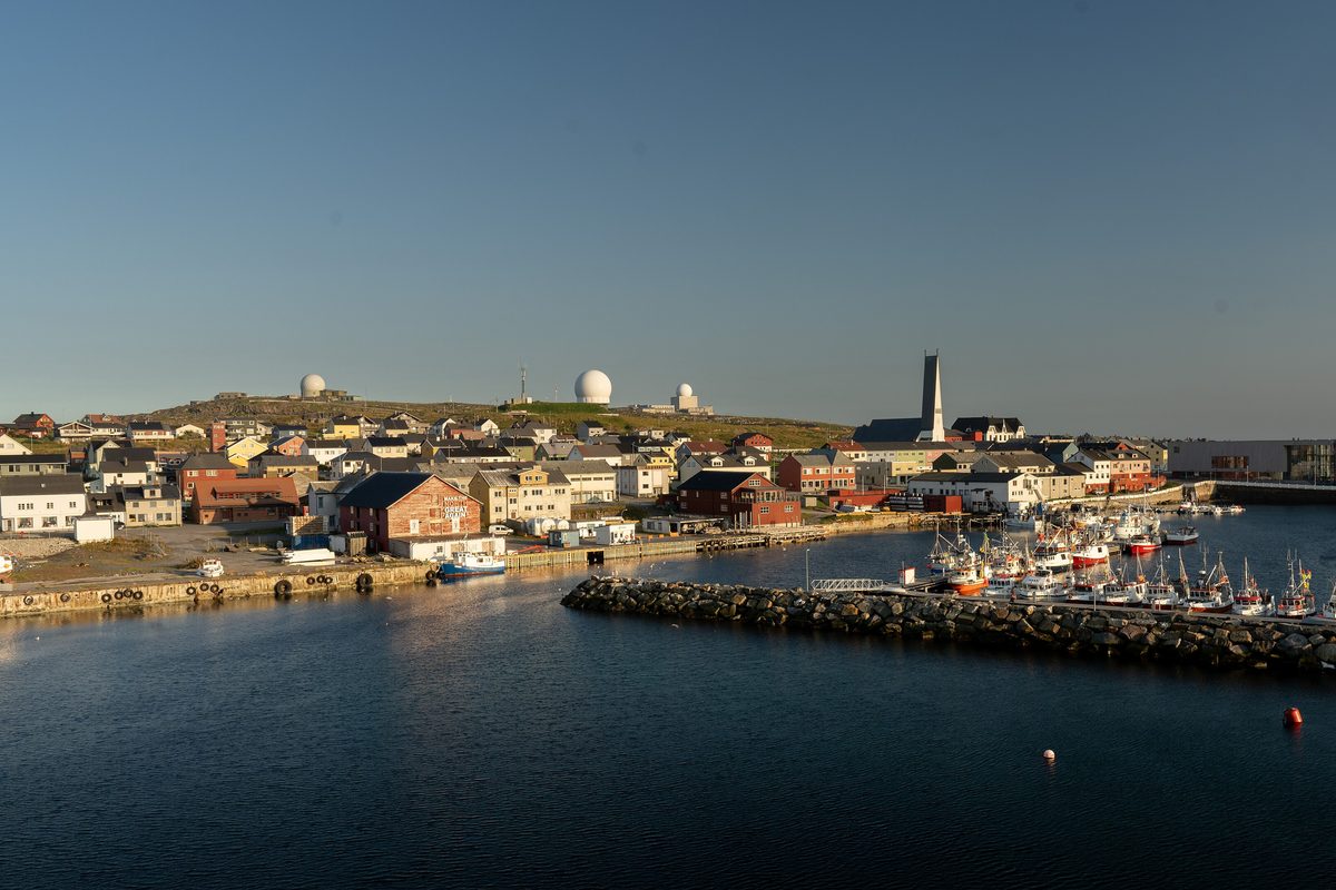 Vardø sett fra sjøen i kveldssol, med havna, fiskebåter, kirketårnet og radarkuplene på høyden bak byen